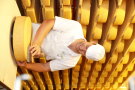 Ein junger Mann kümmert sich um einen großen, runden Laib Käse. Er ist weißgekleidet und trägt eine Kopfbedeckung. Hinter ihm erhebt sich ein Regal mit unzähligen Käselaibern. (Quelle: LfL)