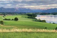 Grüne Wiesen im Vordergrund, rechts ein See und im Hintergrund sind die Berge.