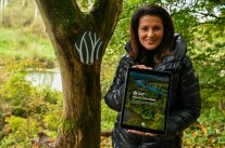 Forstministerin Kaniber steht neben einem Baum mit dem Naturwald-Logo und hält ein Tablet in den Händen