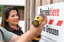 Hütteneigentümer Hans Gruber schraubt mit Ministerin Kaniber das Schild "respektiere deine Grenzen" an eine Scheunenwand.