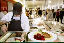 Eine Person mit Schürze und Kappi gibt Essen in einer Kantine aus.