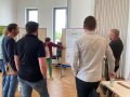 Mehrere Personen stehen im Halbkreis vor einem Flipchart und eine Frau erklärt das Geschriebene