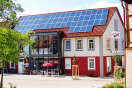 Der Dorfladen mit Mehrgenerationenwerkstatt in Aidhausen. Vor dem Gebäude stehen Stühle, Tische und ein Sonnenschirm. Das Dach des Gebäudes ist mit Photovoltaiksensoren belegt. Links und rechts im Bild steht je ein Baum. Die Fassade des Mehrgenerationenhauses ist im Erdgeschossbereich rot, im Obergeschoss weiß angestrichen.