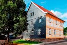 Saniertes zweigeschossiges Fachwerkhaus. Giebelseite grau geschiefert, Längsseite sichtbares, gelbes Fachwerk. Im Vordergrund ein Laubbaum mit halbrunder Sitzban