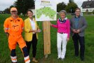 Ein Schild mit der Projektinfo steht in der Mitte, darum stehen vier Menschen.