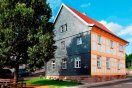 Saniertes zweigeschossiges Fachwerkhaus. Giebelseite grau geschiefert, Längsseite sichtbares, gelbes Fachwerk. Im Vordergrund ein Laubbaum mit halbrunder Sitzbank