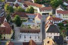 Ortsmitte aus der Vogelperspektive. In der Mitte neuer gestalteter Dorfplatz mit Freifläche und einem Gebäude, in dem Autos parken und auf dessen Dach ein Kinderspielplatz eingerichtet ist.
