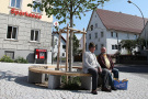 Freifläche im Dorf. Um einen neu gepflanzten Baum ist eine kreisrunde Sitzmöglichkeit angebracht, auf der zwei älteren Männer sitzen und sich unterhalten.