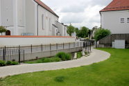 Renaturierter Bachlauf entlang der Einfriedungsmauer einer Kirche. Parallel dazu verläuft eine gepflasterter Fußweg.