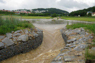 Angestautes Wasser fließt aus einer Wasserrückhaltung (Bodenwelle) kontrolliert durch einen mit Gabionen befestigten Auslass.