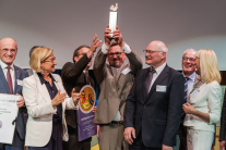 Eine Gruppe feierlich gekleideter Menschen, heben einen Pokal voller Stolz in die Höhe.