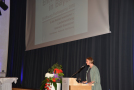 Frau am Rednerpult; Hintergrund Einblendung des Vortragsthemas durch Powerpoint-Präsentation