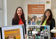 Zwei Frauen hinter zwei Monitoren und für einem Plakat "Soziale Landwirtschaft . eine Perspektive für Ihren Betrien"