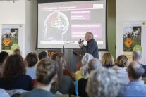 Redner mit Mikrofon, im Hintergrund eine Präsentation auf Leinwand und im Vordergrund die Zuhörer