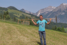 Ein junger Bergbauer posiert mit einem Holzrechen auf den Schultern vor Bergkulisse