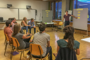 Mehrere Personen sitzen im Halbkreis um einen Moderator, zwei Pinnwände mit gelben Zetteln und eine Flipchart. 