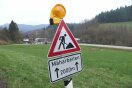 Ein Schild am Straßenrand warnt vor Mäharbeiten. Im Hintergrund ist eine Kreisstraße in einer hügeligen bewaldeten Landschaft zu sehen.