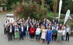 Gruppenfoto mit vielen Menschen im Freien, vorne in der Mitte steht eine Frau mit rotem Jacket.