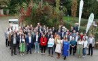 Gruppenfoto mit vielen Menschen im Freien, vorne in der Mitte steht eine Frau mit rotem Jacket.