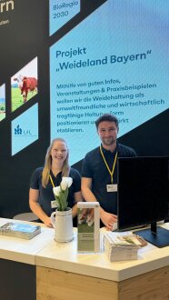 Eine Frau und ein Mann stehen an einer Theke, hinter ihnen läuft eine Präsentation