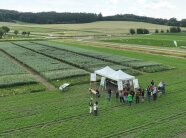 Drohnenaufnahme: Landschaft mit kleineren Versuchsparzellen, im Vordergrund ein Pavillon mit mehrere Menschen.