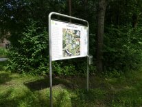 Infotafel an am Wegrand vor Hecken und Bäumen