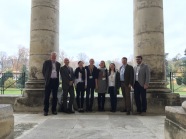 Gruppenfoto von acht Personen, zwischen großen Steinsäulen, im Hintergrund ein Park mit Wiese und Bäumen