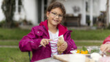 Ein Mädchen sitzt im Garten an einem Tisch und streicht Quark auf ein Stück Stock-Brot.