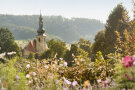 Kirche mit Zwiebelturm, teilweise zu sehen zwischen Bäumen und Blumen.  