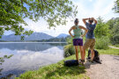 Ein Mann und eine Frau blicken auf einen See im Grünen bei Sonnenschein. Ihre Rucksäcke liegen am Boden.