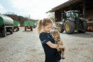 Ein Kind hält eine Katze auf dem Arm, im Hintergrund befinden sich landwirtschaftliche Maschinen.