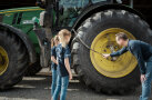Zwei Mädchen blicken auf ein Hinterrad von einem Traktor, an dem ein Mann den Luftdruck prüft.