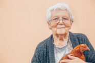 alte Frau mit weißem Haar hält zufrieden schmunzelnd ein Huhn im Arm