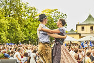 Ein Paar in Dirndl und Lederhose tanzt auf einer Bühne inmitten vieler Menschen.