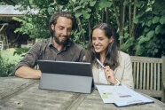 Ein Mann und eine Frau sitzen lachen im Garten an einem Holzisch und sehen gemeinsam in einen Laptop