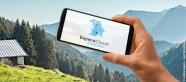 Hand hält ein Handy mit dem BayernCloud App auf dem Bildschirm. Im Hintergrund Berge, Bergwiese und Hütte u