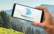 Hand hält ein Handy mit dem BayernCloud App auf dem Bildschirm. Im Hintergrund Berge.