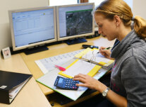 Eine Frau am Schreibtisch vor einem offenen Ordner. Sie bedient einen Taschenrechner und schreibt mit der anderen Hand.