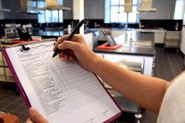 Die Hand einer Frau füllt ein Formular aus, im Hintergrund eine Großküche.