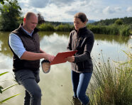 Datenaufnahme: Fischwirt und Wissenschaftliche Mitarbeiterin stehen an Karpfenteich, blicken auf Klemmbrett mit Fragebogen und begutachten Fischfutter.