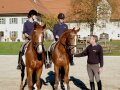 Zwei junge Frauen mit Helm auf dem Kopf sitzen auf zwei braunfarbigen Pferden und erhalten Reitunterricht von ihrem Lehrer.