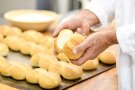 vor einem Blech mit frischgebackener Brötchen und einer mehlbefüllten Schüssel prüft die Bäckerhand eine gerade aufgeschnittene Semmel. (Foto: Tobias Hase, StmELF)
