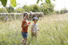 Ein Mädchen und ein Bub stehen inmitten von Gras. Hinter ihnen, in etwas Entfernung sind weidende Schafe zu sehen.