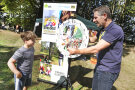 Ein Bub und ein LfL-Mitarbeiter stehen am Wissensrad neben dem Plakat "Streuobst-Schulwochen". Auf dem Rad stehen viele Streuobst-Fragen, die beantwortet werden wollen.