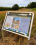 Eine Tafel mit Erklärungen zur Heidelerche steht am Wegesrand des Themenpfades "Biodiversität im Hopfen". Im Hintergrund sind Feld, Hopfen und Wald zu sehen.