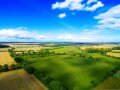 Drohnenbild mit einer grünen Ackerfläche und blauem Himmel mit Wolken. 