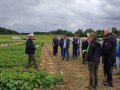 Ein Mann steht in einem Kartoffelacker links mit zwei freigelegten Reihen Kartoffeln vor sich und erklärt einer Gruppe Landwirten, die rechts im Bild stehen,  Neues zum Kartoffelanbau.