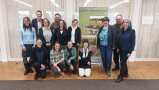 Zehn Frauen und drei Männer stehen und knien fürs Gruppenfoto vorm Plakat zur Wiesenmeisterschaft Ostallgäu.