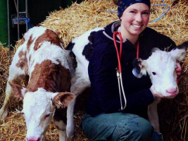 Auf einem Strohhaufen sitzt eine lachende Frau. Rechts und links von ihr je ein Kalb.