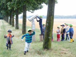 Kinder stehen vor einem überdimensionalen Fernrohr am Feldrand.